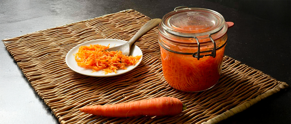 Carottes lactofermentées un bocal en verre et une petite assiette contenant des carottes rapées fermentées, avec une carotte entière à côté